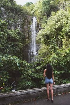 a woman standing in front of a waterfall looking down at the ground with trees around her