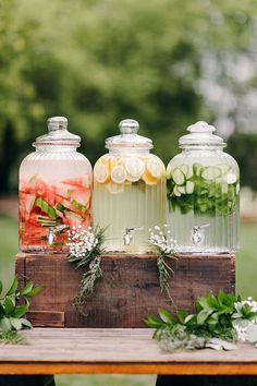three glass jars filled with different types of drinks on top of a wooden table next to flowers and greenery