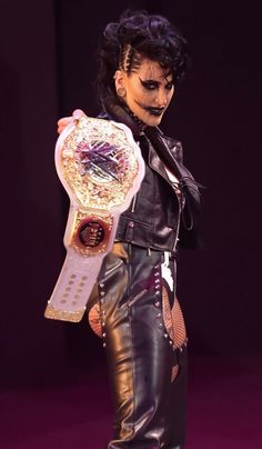a woman in leather pants and makeup holding a wrestling ring on her right hand while standing on a purple surface