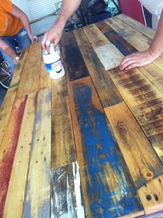 two people sitting at a wooden table with paint on it and one person painting the wood