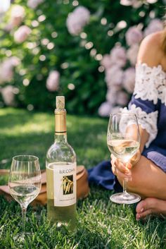 a woman sitting in the grass next to two wine glasses and a bottle of wine