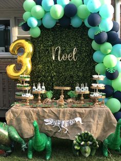 a table topped with balloons and desserts next to a wall covered in greenery