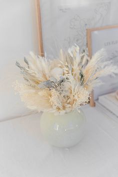 a white vase filled with dried flowers on top of a table next to framed pictures