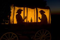 two people sitting at a table in front of a covered wagon with bottles on it