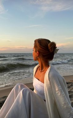 a woman sitting on top of a sandy beach next to the ocean in white clothing