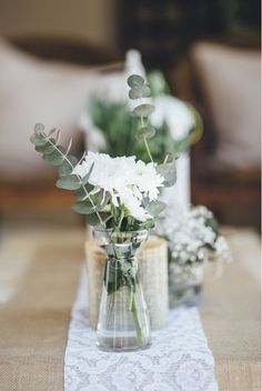 two vases filled with white flowers sitting on top of a tablecloth covered table