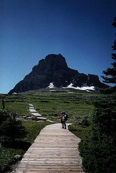 two people walking down a wooden path towards a mountain