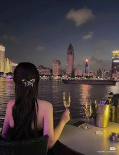 a woman sitting at a table with two wine glasses in front of her, overlooking the city skyline