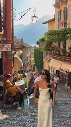 a woman in a white dress is walking down the cobblestone street with people sitting at tables