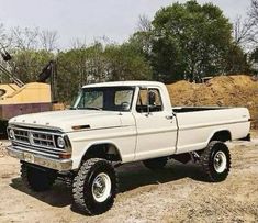 an old white pickup truck parked in the dirt near a pile of sand and trees