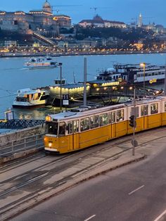 a yellow and white train traveling down tracks next to a body of water with boats in the background