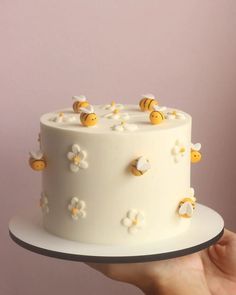 a hand holding a white cake with yellow bees on top and flowers around the edges