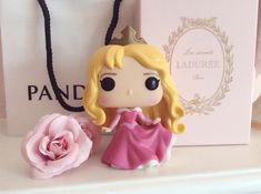 a little princess figurine sitting next to a pink rose on a shelf with a gift bag in the background