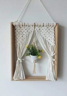 a white wall hanging with some flowers in the window and curtains around it, as well as a potted plant