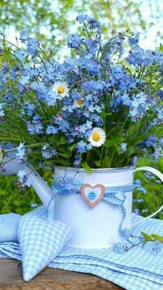 a watering can filled with blue flowers sitting on top of a wooden table next to a gingham cloth