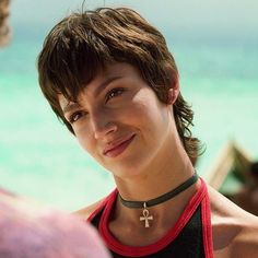 a woman with a cross on her neck smiling at another person in the background and water behind her