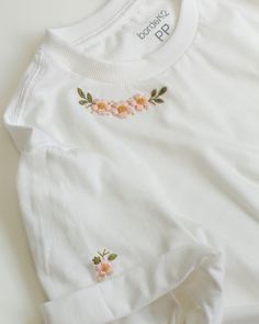 a white t - shirt with flowers embroidered on the chest and sleeves, sitting on top of a table