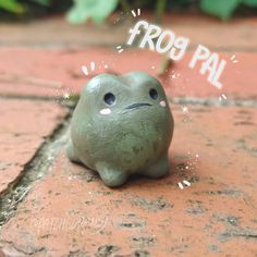 a small green frog toy sitting on top of a brick floor with the words frog pal written above it