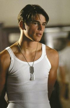 a young man wearing a white tank top with a silver necklace on his neck is looking at the camera