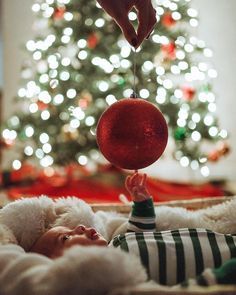 a baby is laying down in front of a christmas tree with a red ball hanging from it's head