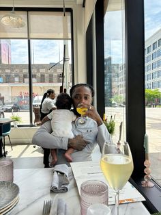 a woman holding a baby while sitting at a table in front of a wine glass