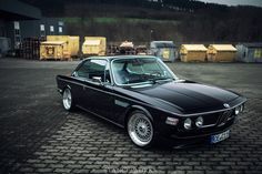 an old black car parked in front of some shipping containers on a brick floored area