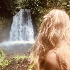 a woman is looking at a waterfall in the woods with her hair blowing in the wind