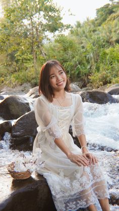 a woman sitting on top of a rock next to a river wearing a white dress