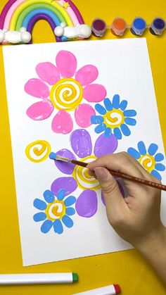 a child's hand holding a paintbrush and painting flowers on paper with rainbows in the background
