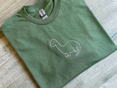 a green t - shirt with a white dinosaur embroidered on the front and back, sitting on a wood floor