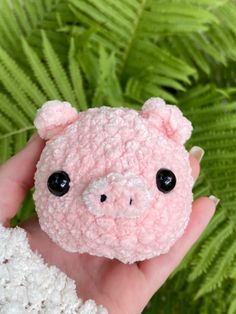 a pink crocheted teddy bear with black eyes is held in front of a fern