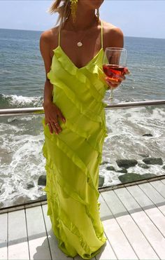 a woman in a yellow dress holding a glass of wine and looking at the ocean