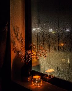 a window sill with candles and flowers on it in front of a rain soaked window