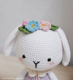 a crocheted bunny with flowers on its head sitting on a wooden table in front of a gray wall