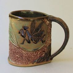 a brown and green coffee mug with two bugs on it's side, sitting on a white surface