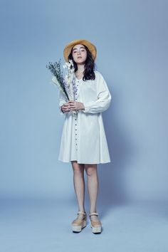 a woman in a white dress and straw hat holds flowers while standing against a blue background