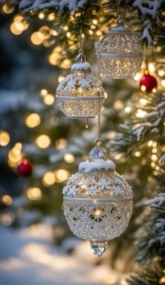 three glass ornaments hanging from a christmas tree with lights in the background and snow on the branches