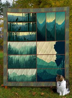 a cat sitting in the grass next to a quilt with trees and mountains on it