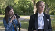 two women standing next to each other in front of trees and one holding a camera