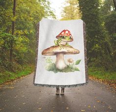 a woman holding up a mushroom blanket in the middle of a road with trees behind her