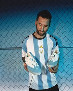 a man holding two pairs of shoes in front of a chain link fence with blue and white stripes