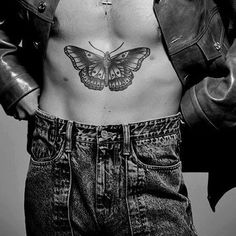 a shirtless man with a butterfly tattoo on his chest and jeans, standing in front of a wall
