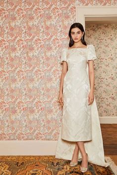 a woman standing in front of a floral wallpapered background wearing a white dress