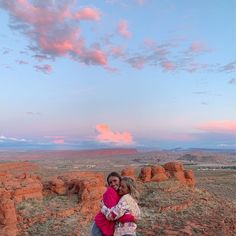 two people hugging each other in front of some rocks and sky with clouds above them