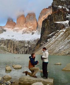 two men are standing on rocks in front of a mountain lake and snow covered mountains