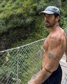 a shirtless man standing on a bridge with trees in the background and bushes behind him