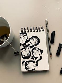 a cup of tea next to a notebook with the faces of people drawn on it