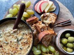 an assortment of food including bread, grapes and apples on a wooden platter with two bowls of dip