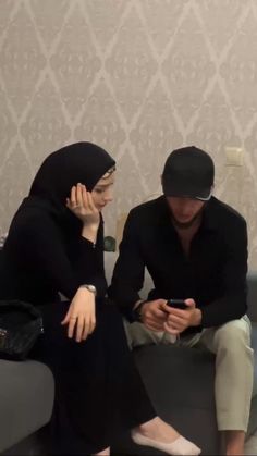 two people sitting on a couch looking at their cell phones while one person is wearing a headscarf