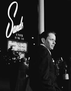 black and white photograph of man standing in front of sands casino sign at night time